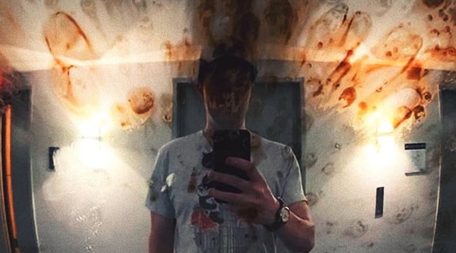 CLOUDKICKER Unveils New Track “The Whirl” The Mosh Network - Ben Sharp taking a mirror selfie in a dimly lit room, with the mirror covered in smudges and reddish stains that create a distorted, eerie effect.
