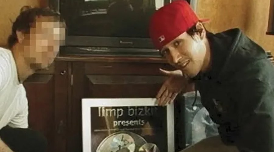 Buddyhead’s Travis Keller Recalls Stealing Fred Durst’s Red Hats and the Fallout That Followed The Mosh Network - A man in a red cap points to a framed Limp Bizkit plaque while posing next to another person with a pixelated face, in a room with a TV and speakers in the background.