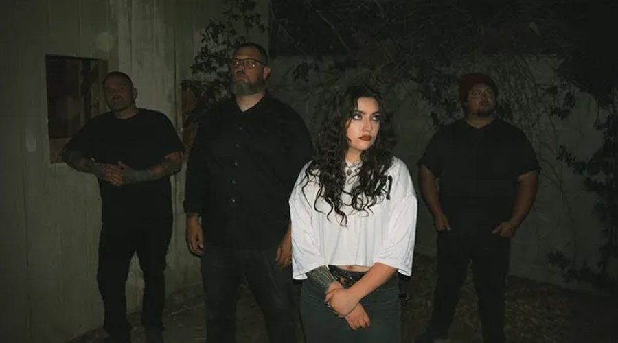 ROMAN CANDLE Sign With Sumerian Records, Release New Single “Fire In The Night Sky Forever” The Mosh Network - Promo photo of the Las Vegas screamo band Roman Candle standing outdoors at night. Four members pose against a dimly lit wall surrounded by trees and foliage. The female vocalist stands in front, wearing a white shirt, while three male members stand behind her in dark clothing.